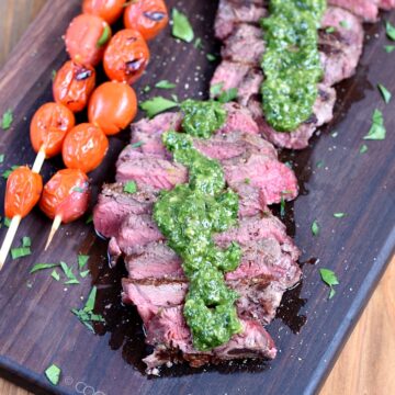 Sliced Sirloin Steak topped with Chimichurri sauce on a wooden cutting board with skewered tomatoes on the side.