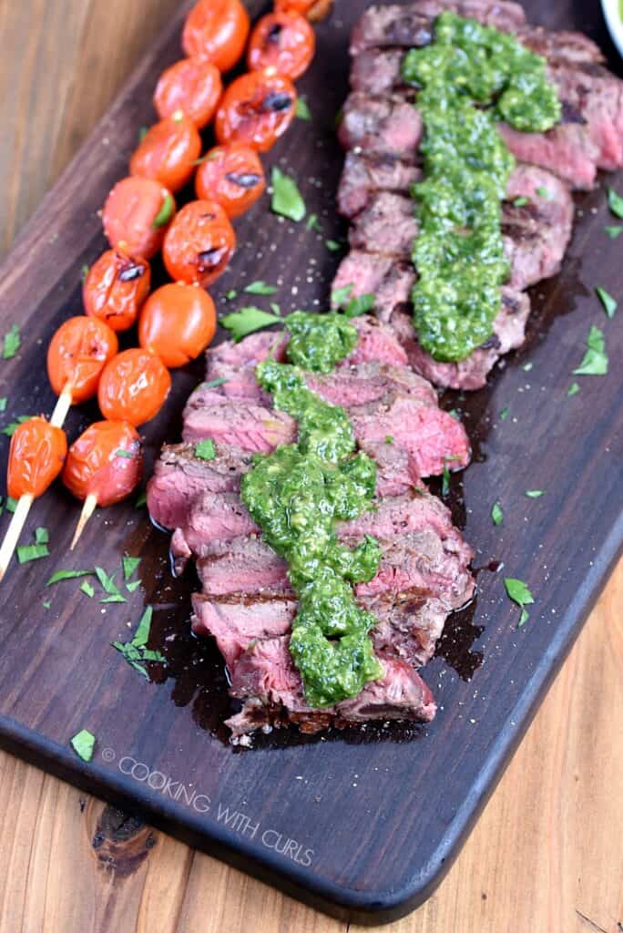 Sliced Sirloin Steak topped with Chimichurri sauce on a wooden cutting board with skewered tomatoes on the side.