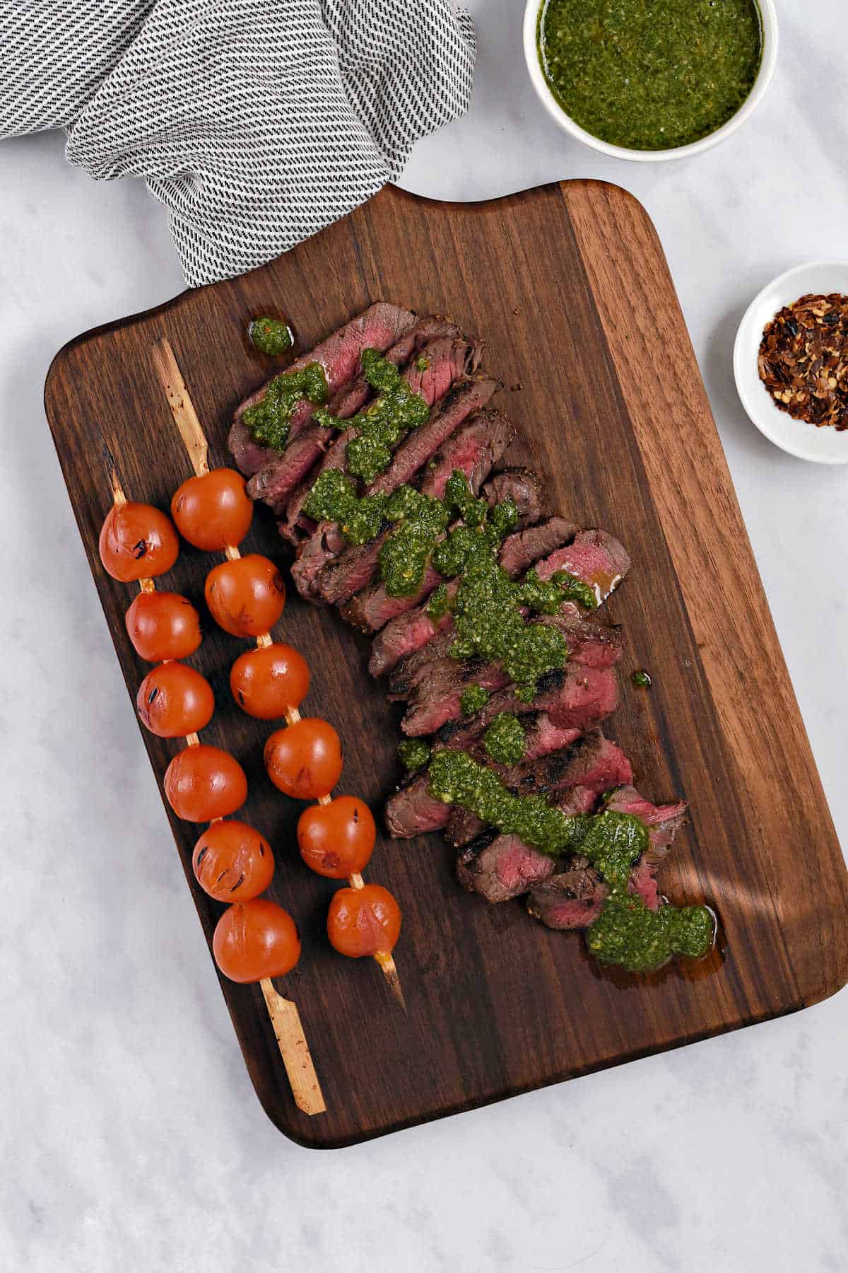 Sliced steak topped with chimichurri sauce on a wooden serving board with two skewers of grilled cherry tomatoes on the side.