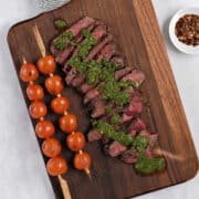 Sliced steak topped with chimichurri sauce on a wooden serving board with skewered tomatoes on the side.