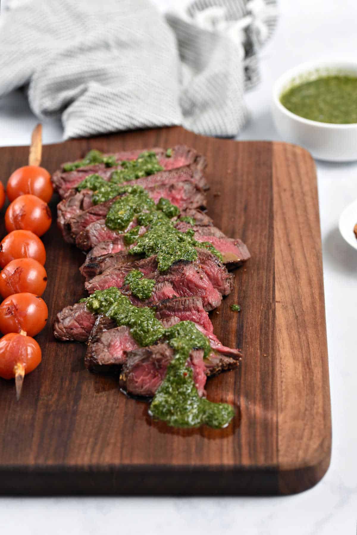 Sliced steak topped with chimichurri sauce on a wooden serving board with skewered tomatoes on the side.