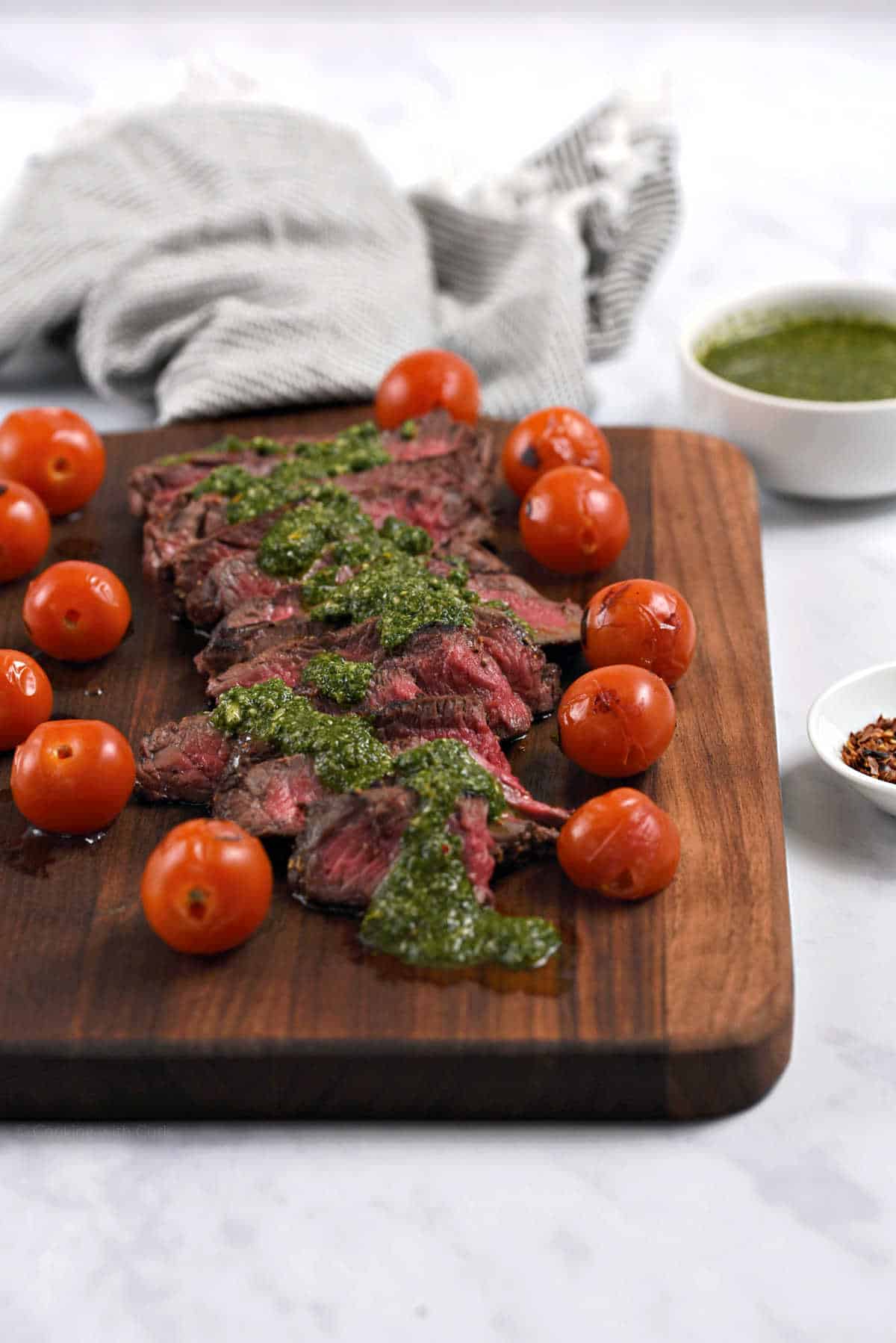 Sliced steak topped with green sauce on a wooden board with burst cherry tomatoes on the edges.