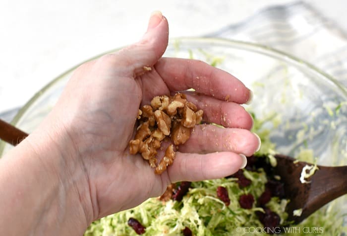 A hand crushing walnuts over a glass bowl of shaved Brussels sprouts.