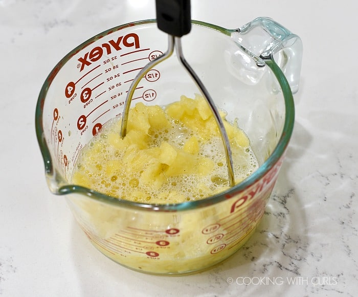 Fresh pineapple in a large measuring cup being pressed with a potato masher 