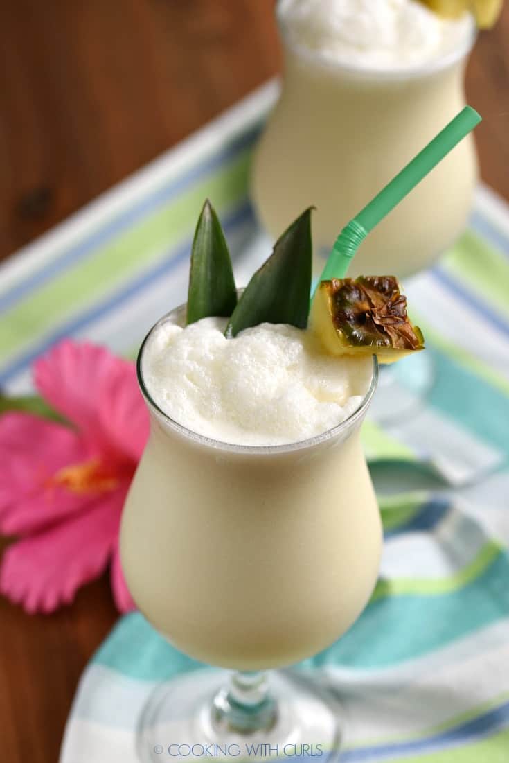 Two traditional pina colada cocktails garnished with a pineapple wedge, straw and pineapple leaves sitting on a striped napkin