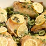 Close up image of chicken thighs topped with olives, capers, lemon slices and thyme