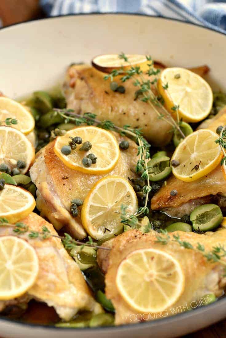 A close up image of chicken thighs topped with olives, capers and lemon slices in a white skillet