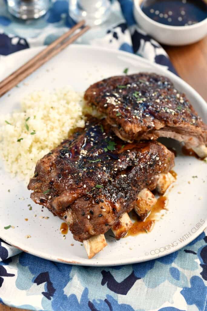 Two stacks of pork ribs on a plate next to cauliflower rice with a small bowl of teriyaki sauce in the corner