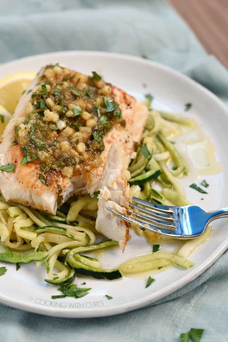 A flaky bite of mahi mahi on a fork with the remaining fillet in the background on a bed of zucchini noodles