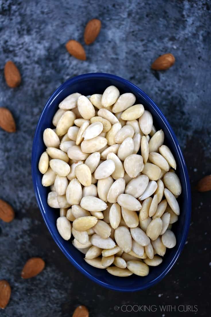 An oblong blue bowl filled with blanched almonds with raw almonds in the background