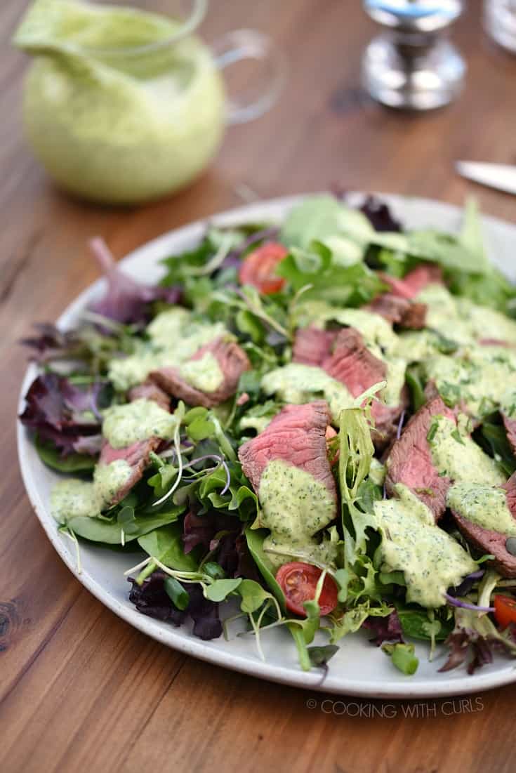 Cilantro-Lime dressing topped steak salad on a large white plate with a carafe of dressing in the background.