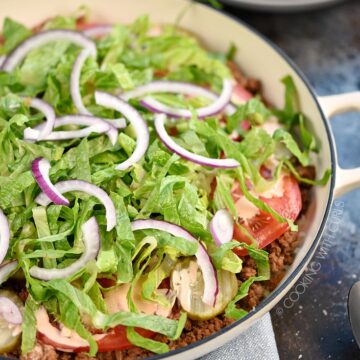 A skillet filled with ground beef, tomato and pickle slices, shredded lettuce and thinly sliced onions