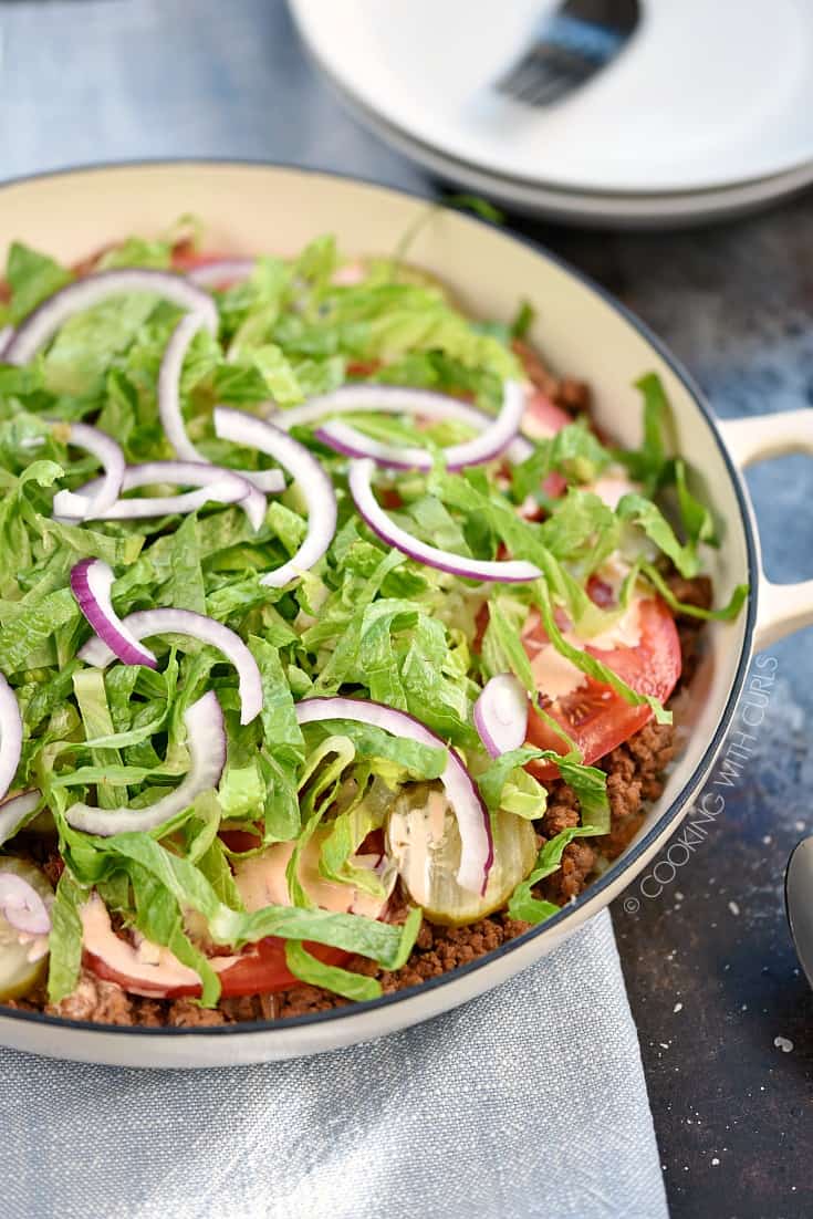 A skillet filled with ground beef, tomato and pickle slices, shredded lettuce and thinly sliced onions