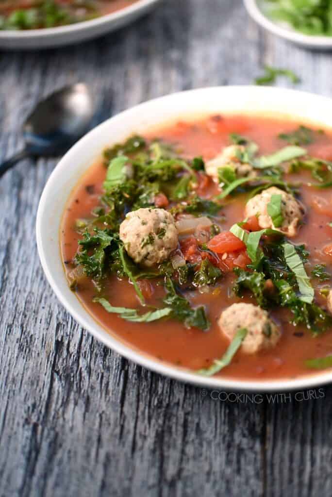 A bowl of Italian Meatball Soup with a second bowl of soup and a dish of basil in the background