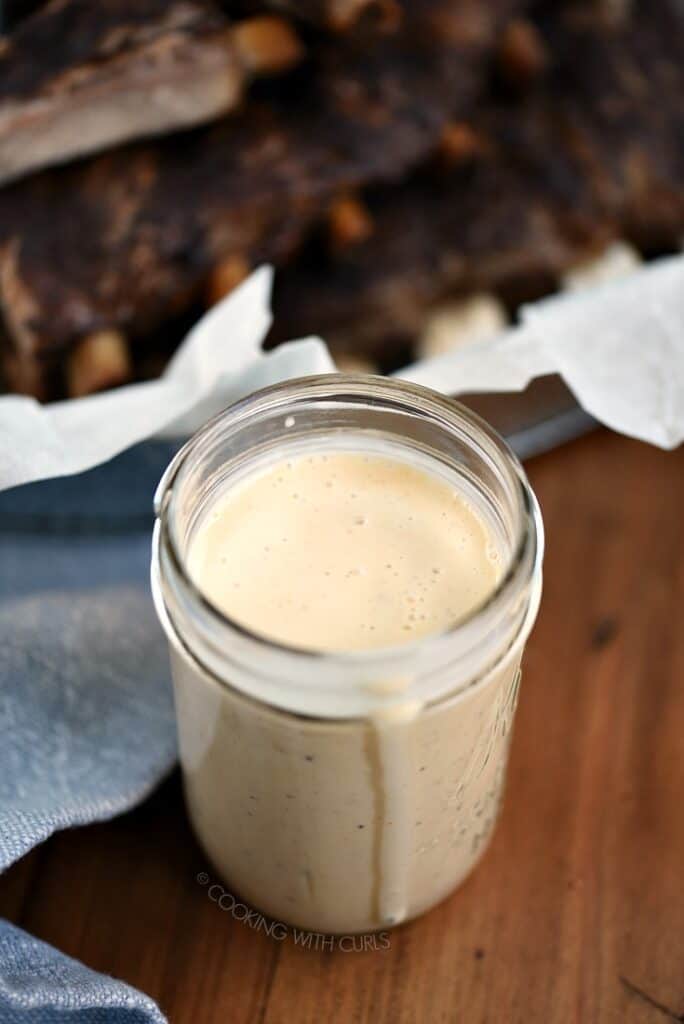 A jar filled with Alabama White Barbecue Sauce with a basket of ribs in the background.