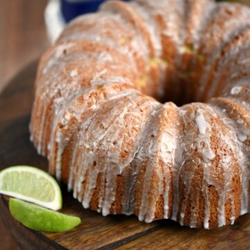 A Margarita Bundt Cake displayed on a wooden cake plate with lime slices on the side.
