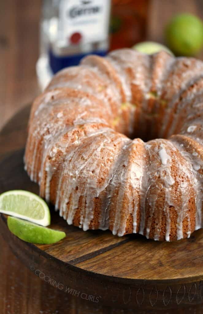 A Margarita Bundt Cake displayed on a wooden cake plate with lime slices on the side.