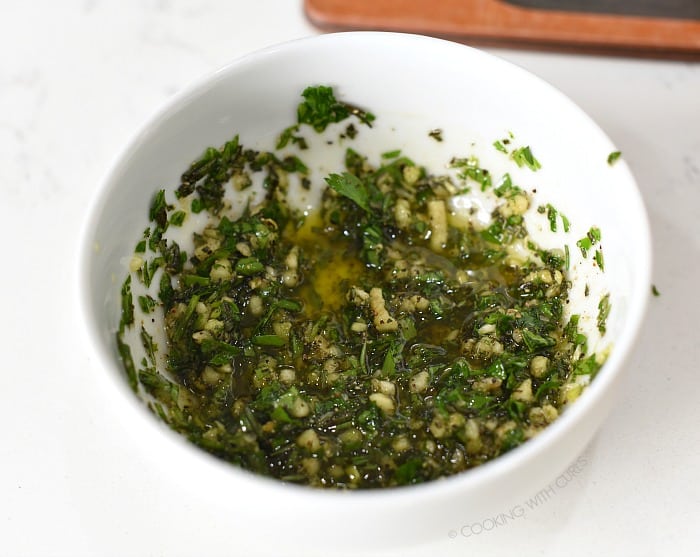 Chopped rosemary, garlic, parsley salt and pepper combined with oil in a small white bowl. 