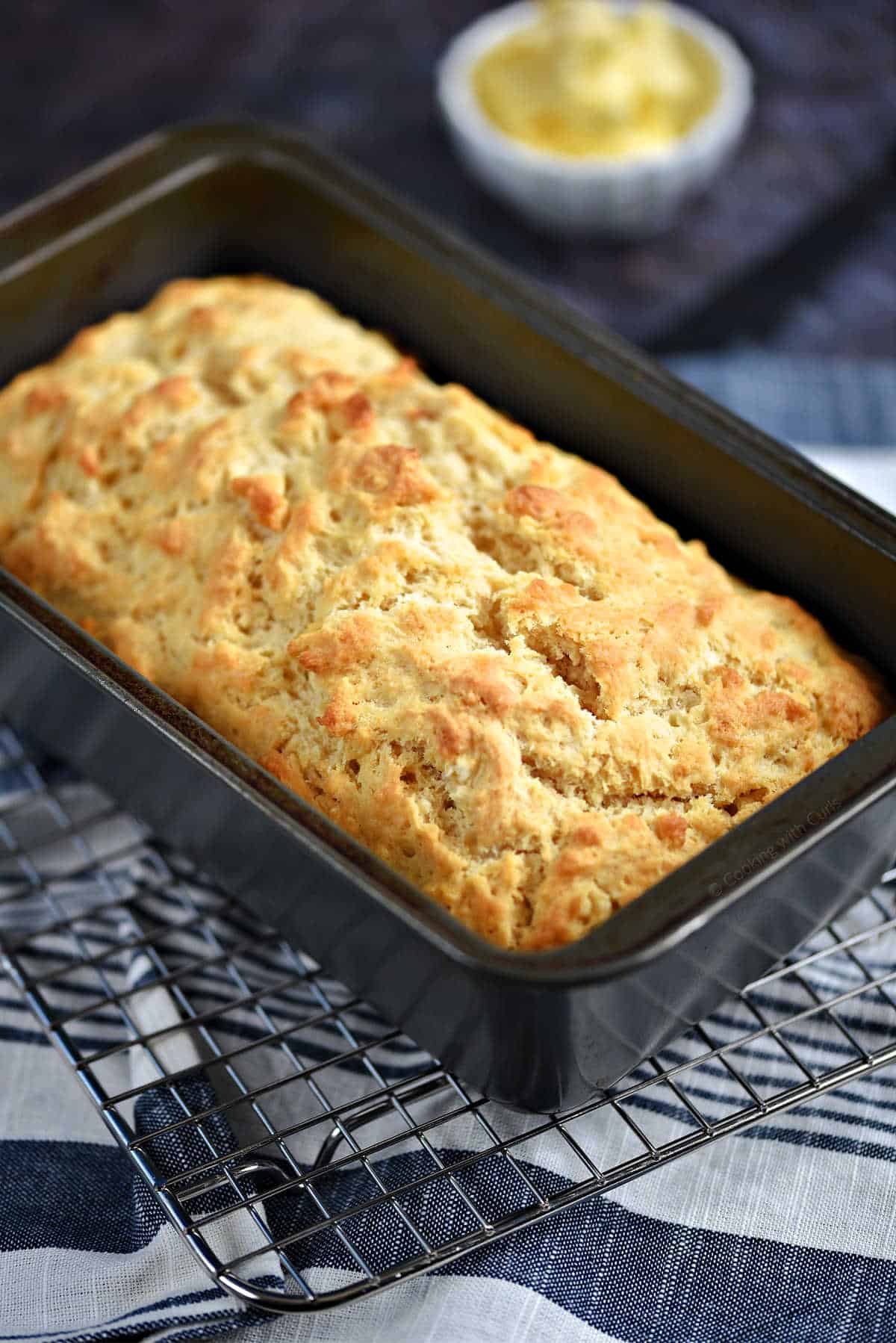 A golden brown loaf of beer bread in a loaf pan sitting on a wire rack.