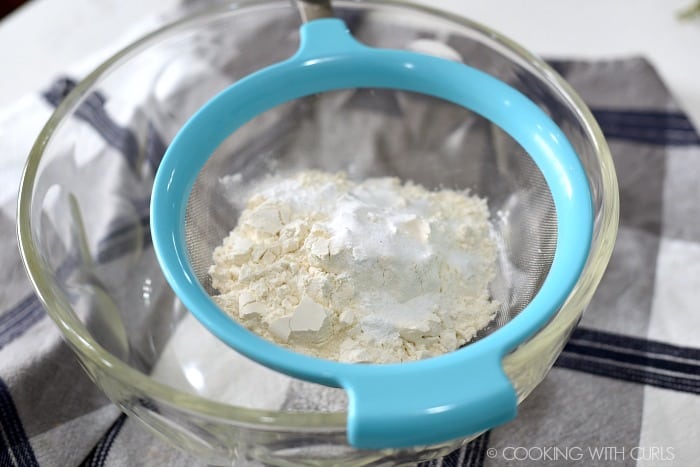 Flour, baking powder and salt in a sifter resting on a glass bowl. 