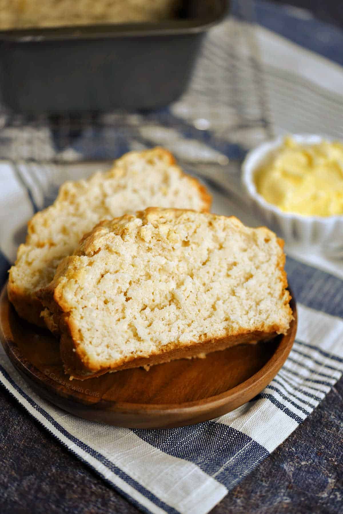 Slices of beer bread on a wooden plate with a bowl of butter in the background.