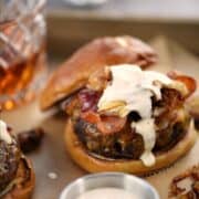 A burger topped with grilled onions, bacon, and whiskey mayonnaise on a pretzel bun with a glass of whiskey in the background with title graphic across the top.