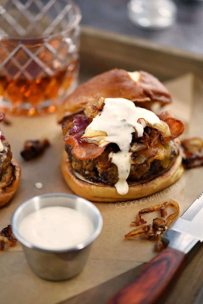 A burger topped with grilled onions, bacon, and whiskey mayonnaise on a pretzel bun with a glass of whiskey in the background.