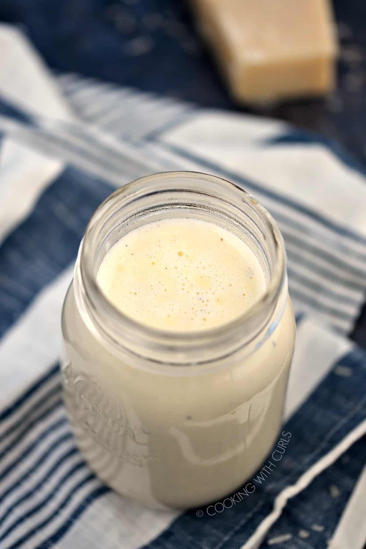 A glass mason jar filled with Classic Alfredo Sauce sitting on a blue and white stripped napkin with a wedge of Parmesan cheese in the upper corner.