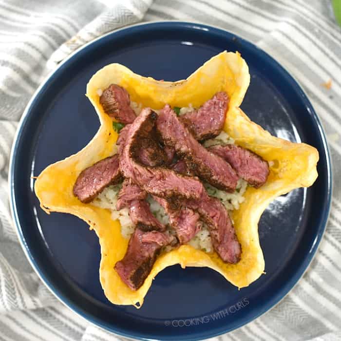 Melted cheese topped tortilla shell with strips of grilled steak on top of the rice sitting on a dark blue plate. 