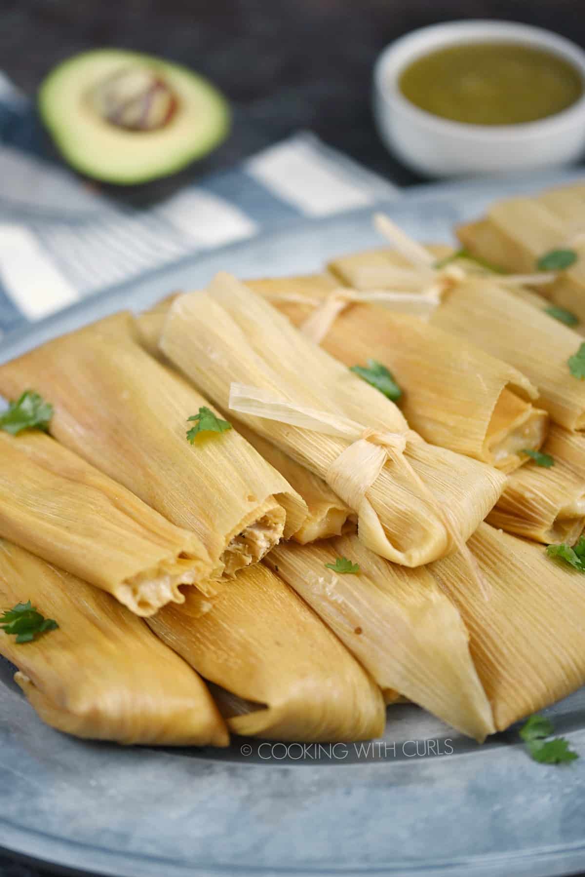Instant Pot Salsa Verde Chicken Tamales stacked on a silver platter with a bowl of salsa verde and a cut avocado in the background.