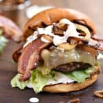 Grilled Double Bacon Burgers with lettuce, tomato, caramelized onions, white cheddar, strips of bacon and white barbecue sauce sitting on a wooden board.