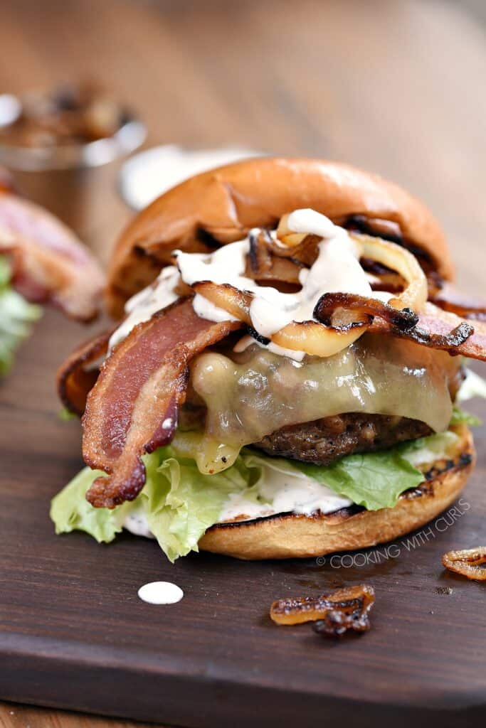 Grilled Double Bacon Burgers with lettuce, tomato, caramelized onions, white cheddar, strips of bacon and white barbecue sauce sitting on a wooden board.