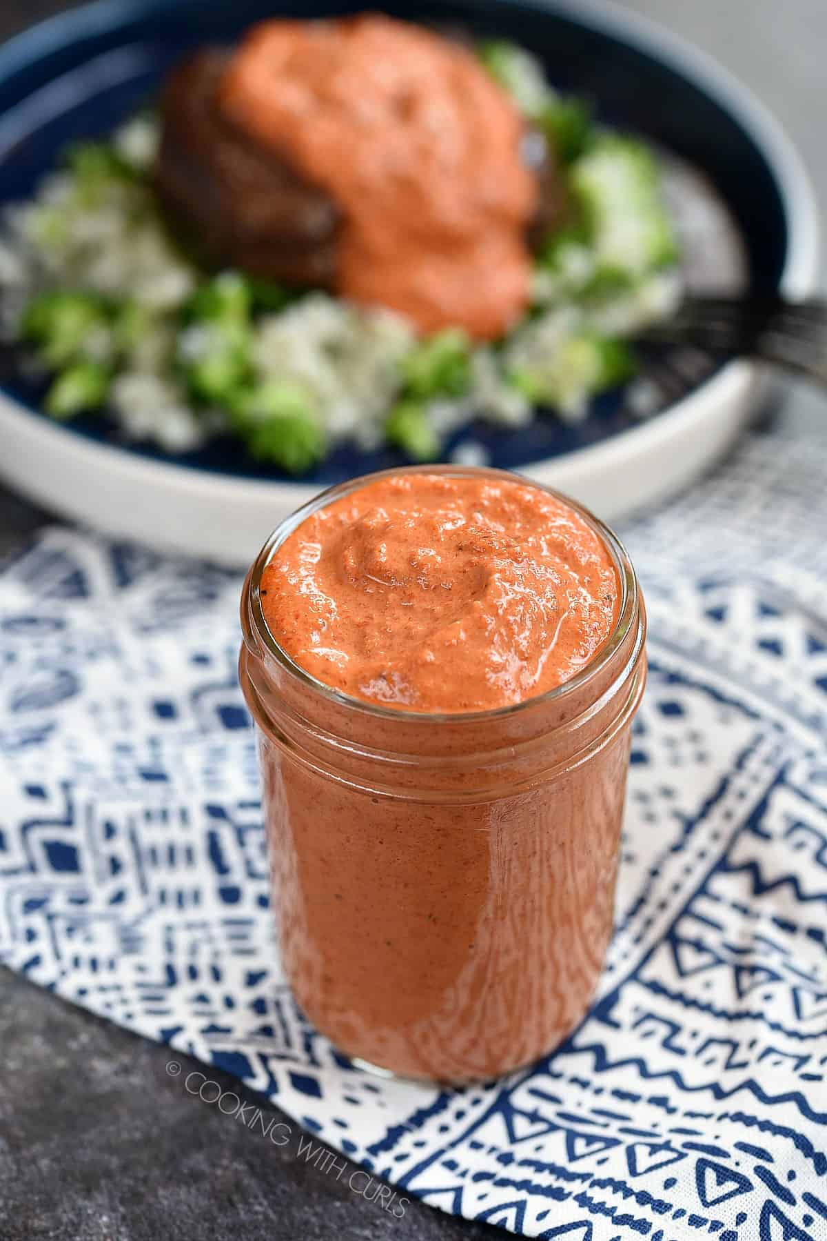 A glass jar filled with roasted Red Pepper Pesto Sauce sitting on a blue and white patterned napkin with a sauce covered steak on a blue plate in the background.