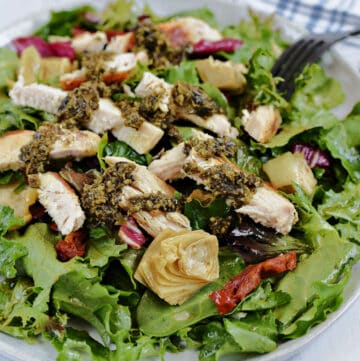 Mixed salad greens tossed with artichoke hearts, sun-dried tomatoes and a light dressing with sliced chicken drizzled with pesto on a large white plate.