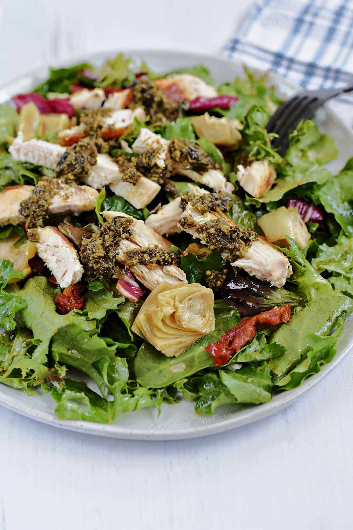 Mixed salad greens tossed with artichoke hearts, sun-dried tomatoes and a light dressing with sliced chicken drizzled with pesto on a large white plate.