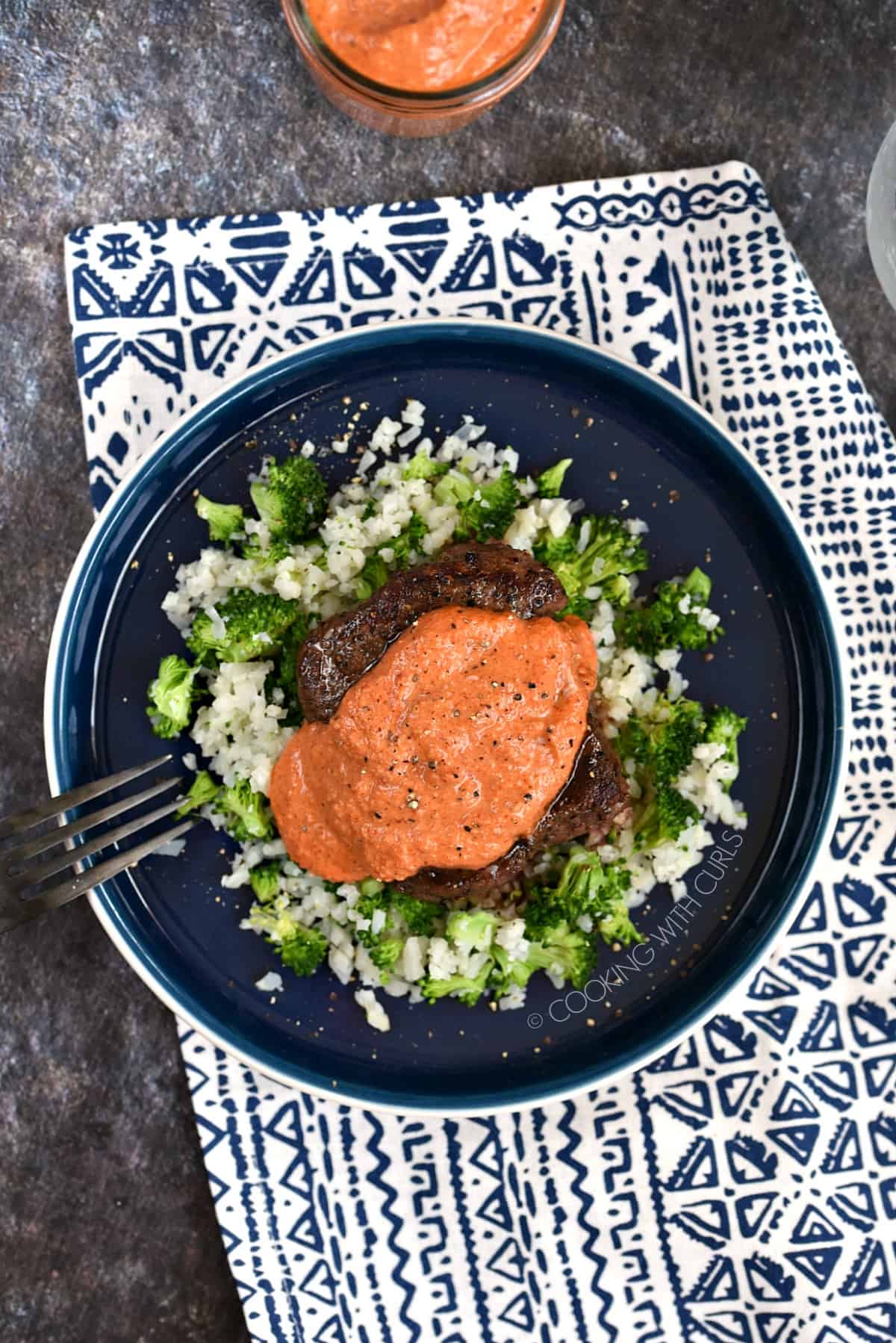 Looking down on a roasted Red Pepper Pesto Sauce topped steak on a bed of cauliflower and broccoli rice sitting on a blue plate.