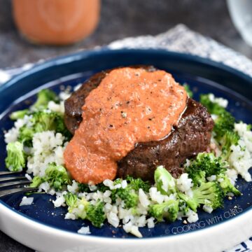 A steak sitting on top of a bed of cauliflower and broccoli rice topped with red pepper peasto sauce.