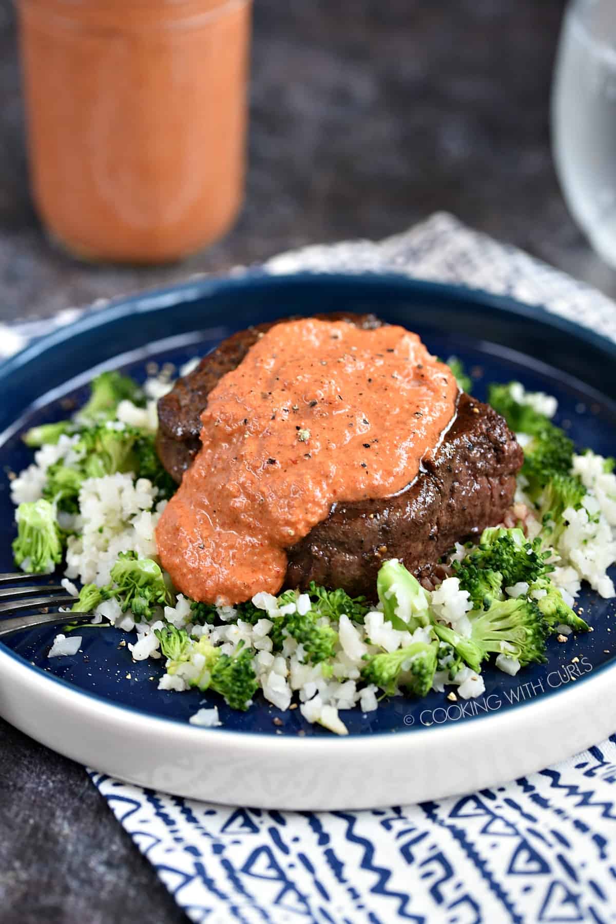 A steak sitting on top of a bed of cauliflower and broccoli rice topped with red pepper peasto sauce.