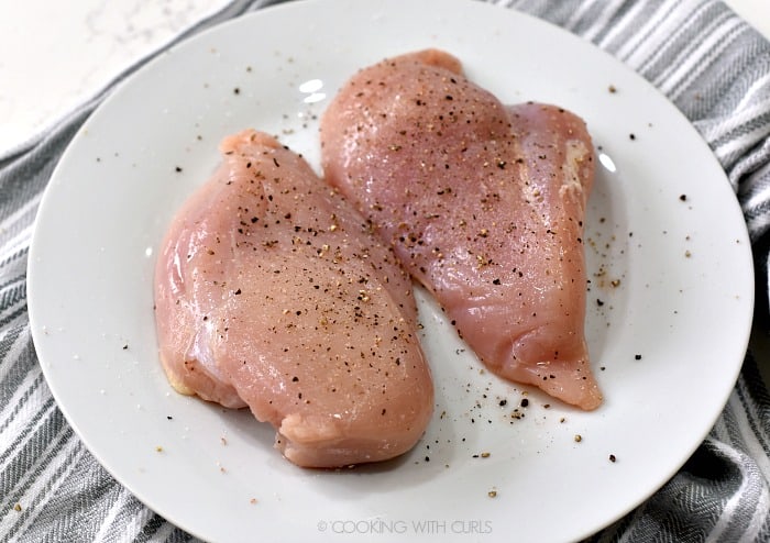 Two boneless, skinless chicken breasts on a white plate sprinkled with salt and pepper.