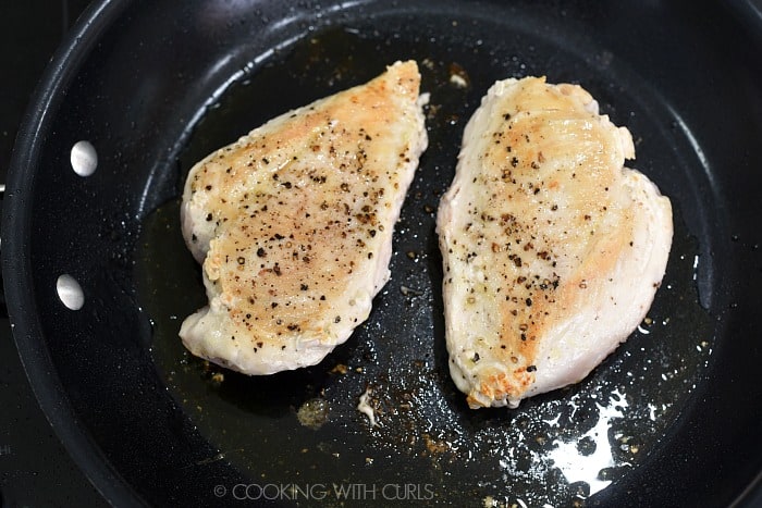Two salt and pepper coated boneless, skinless chicken breasts browning in a non-stick skillet.