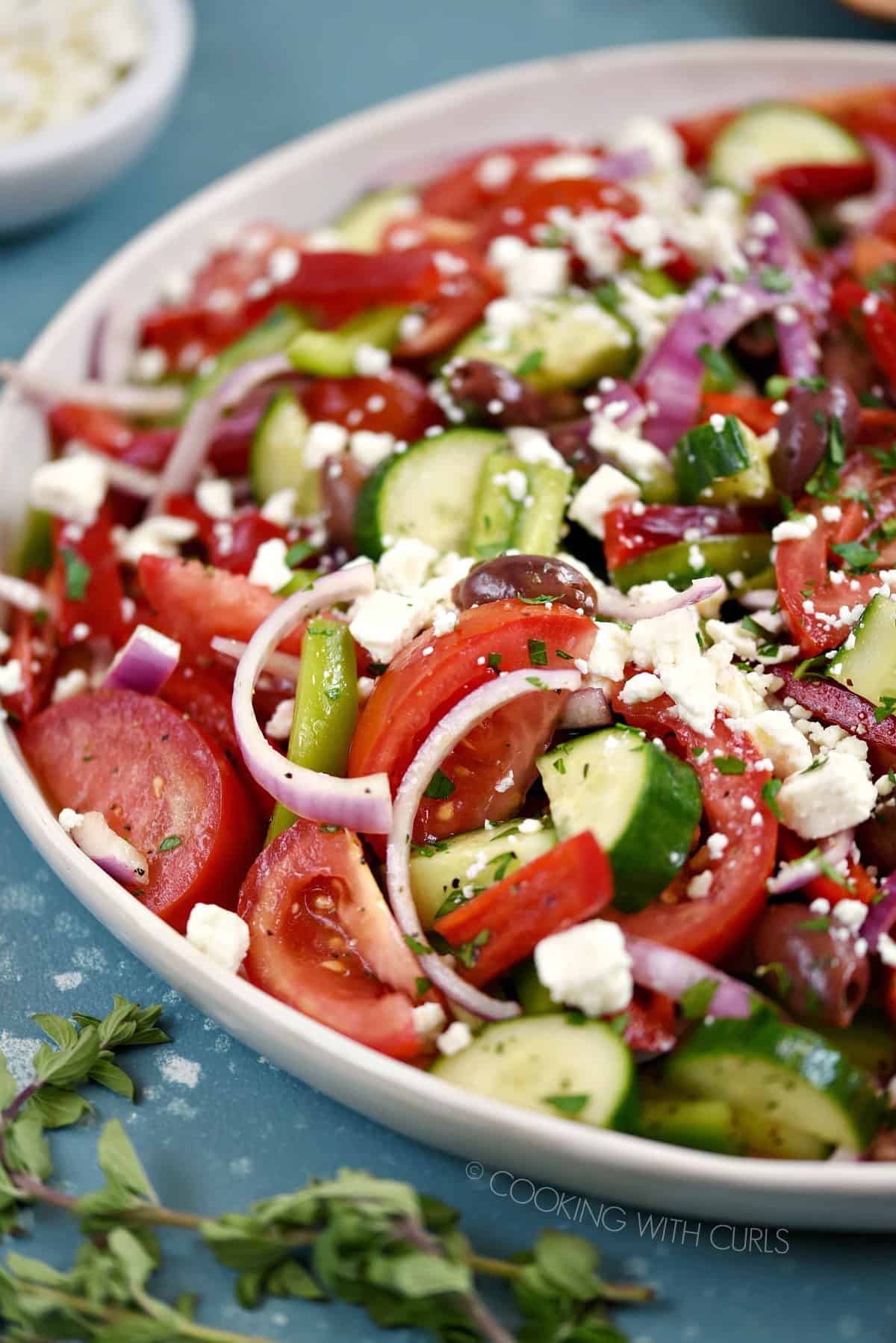 A close-up of tomato wedges, onion, cucumber and bell pepper slices topped with a light vinaigrette and feta cheese in a large white serving platter.