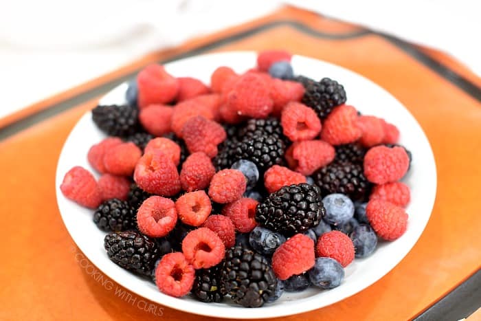 Blackberries, blueberries and strawberries in a white bowl sitting on a wood cutting board. 