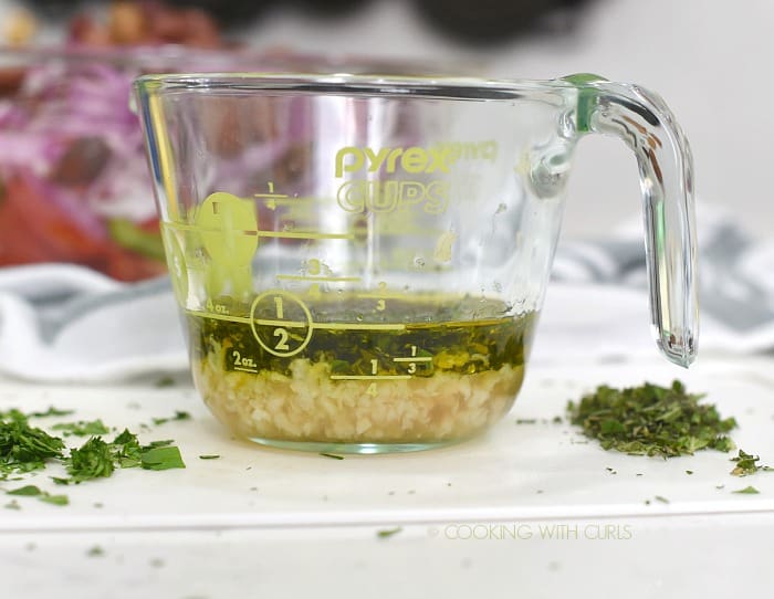 Chopped garlic, parsley, oregano, lemon juice, olive oil and vinegar in a small glass measuring cup. 