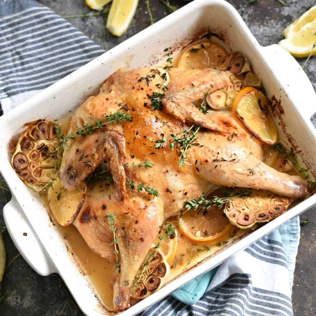 spatchcock chicken with garlic and lemon slices in a white baking dish.