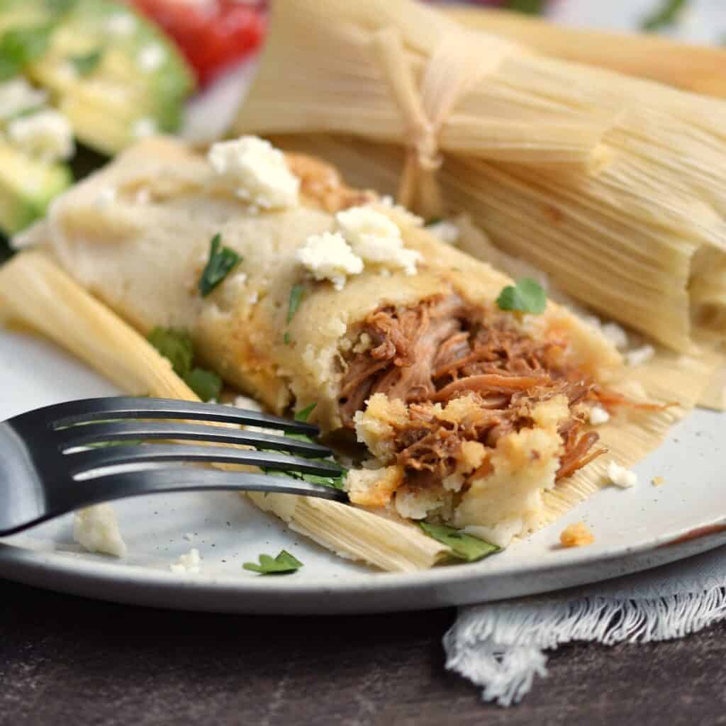 A close-up look at a pork tamale that has been cut into with a black fork, sitting next to a wrapped tamale.