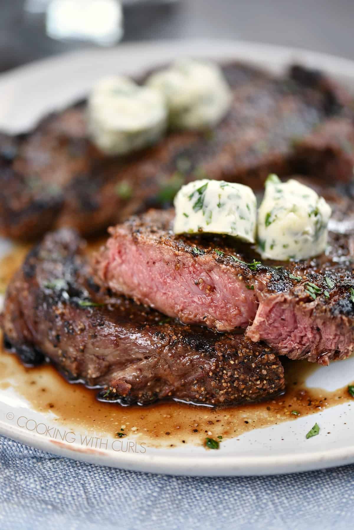Two Perfect Grilled Steaks topped with herb butter with one cut into to show a medium-rare center.