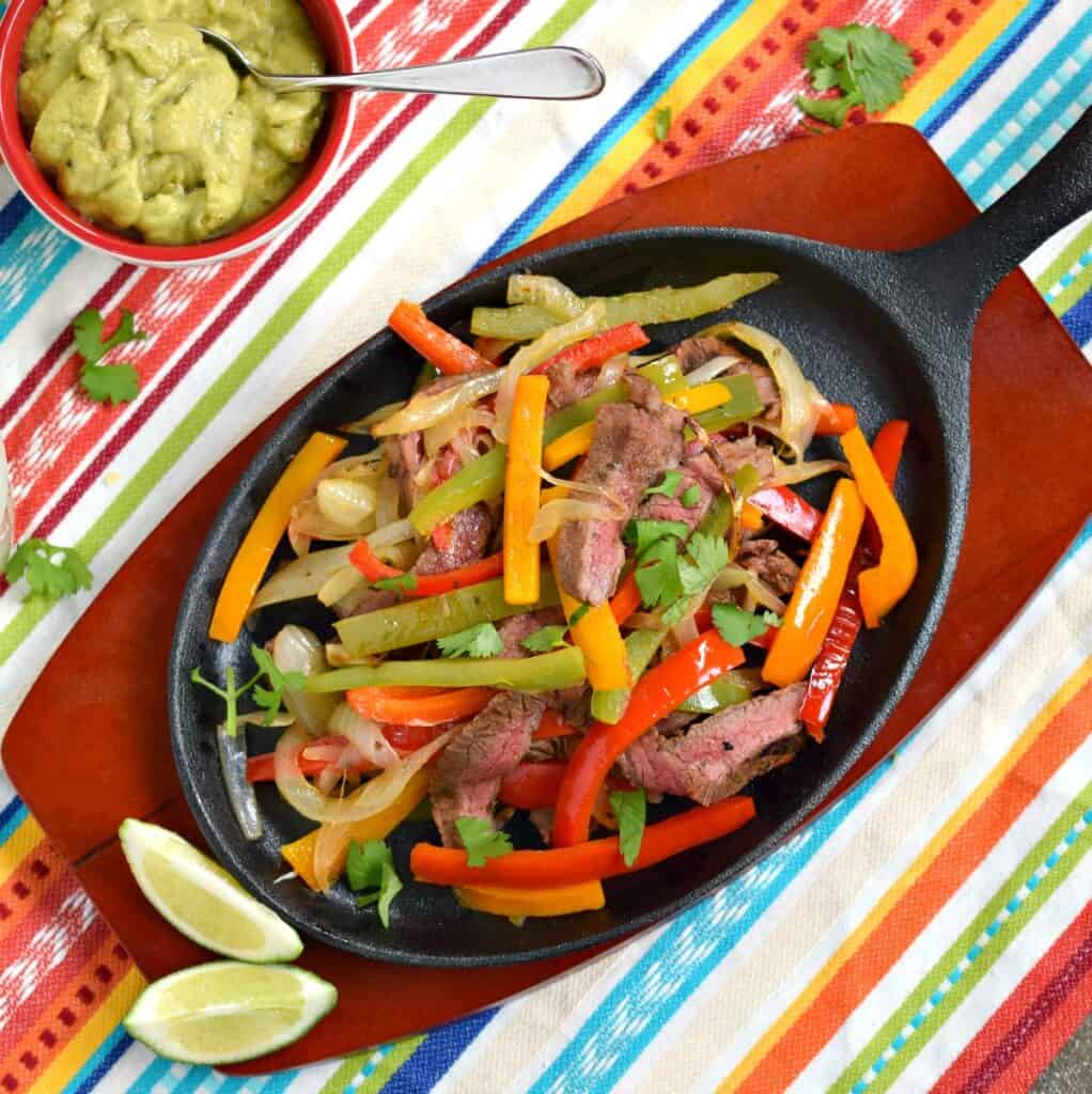 steak, onion and pepper slices cooked in an oval skillet with a bowl of guacamole on the side.
