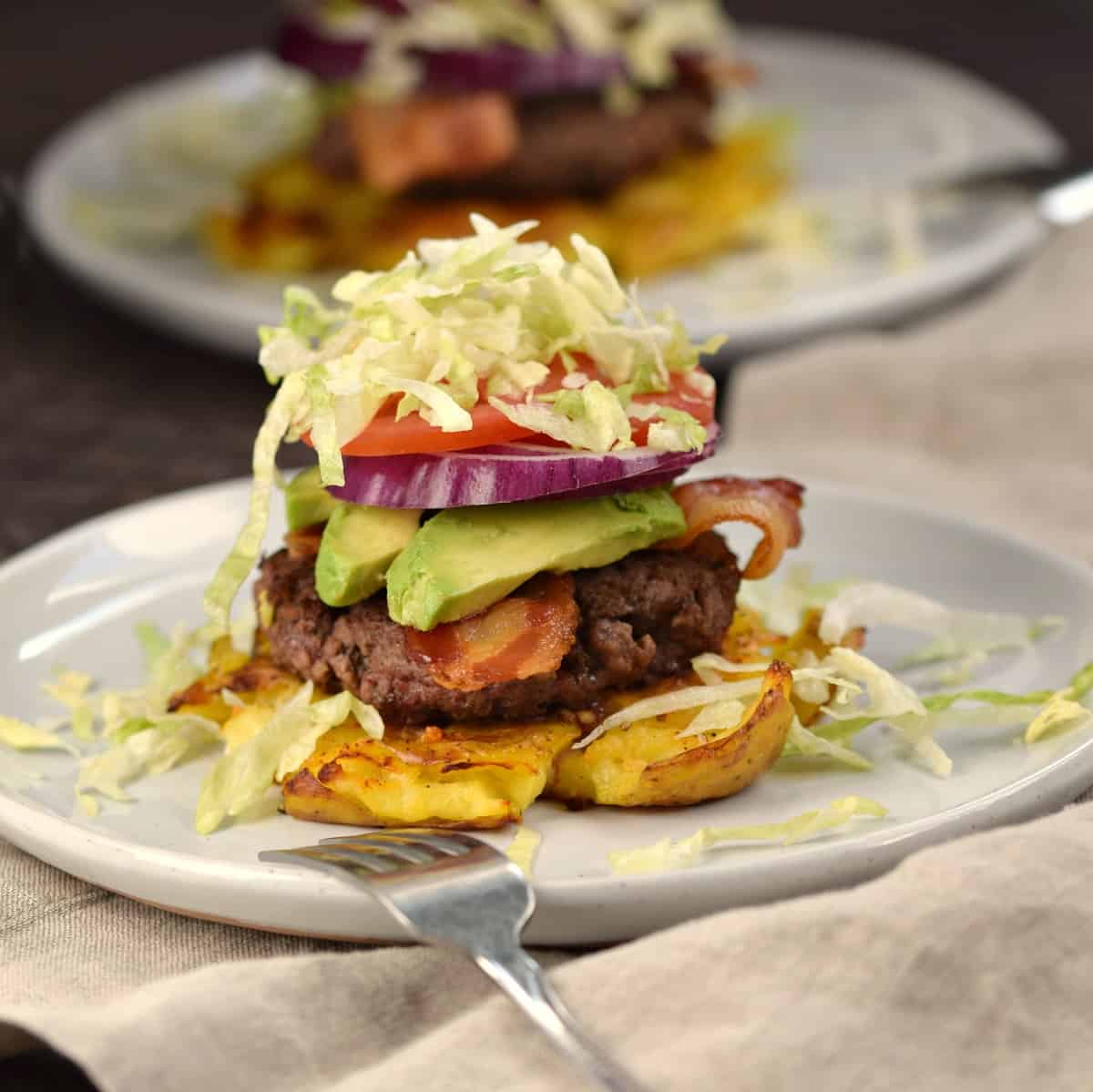 A smashed Yukon gold potato topped with a burger patty, bacon, avocado, onion and tomato slices with shredded lettuce on top.