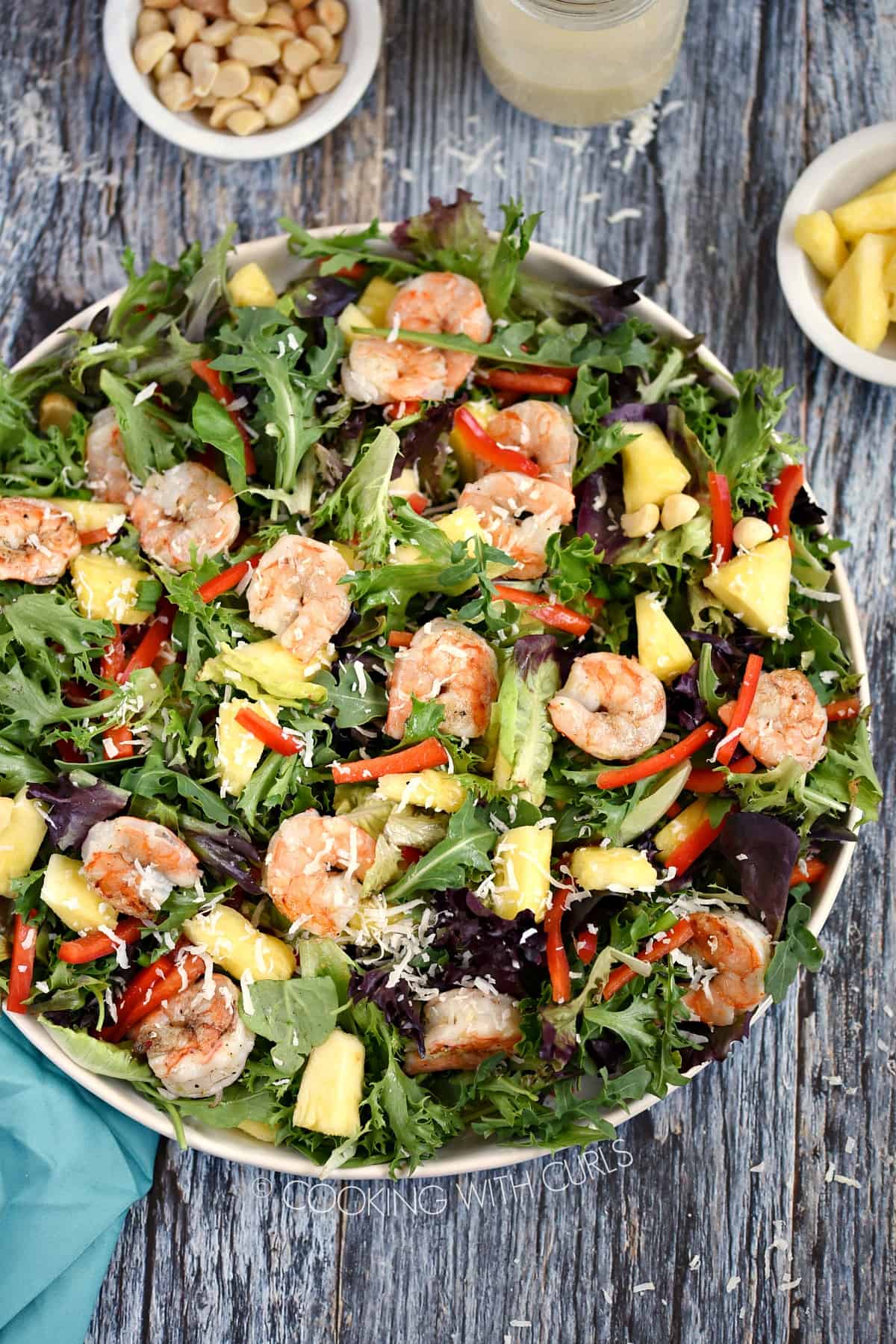Looking down on a white platter filled with Pina Colada Grilled Shrimp Salad with bowls of macadamia nuts and pineapple chunks across the top of the image.