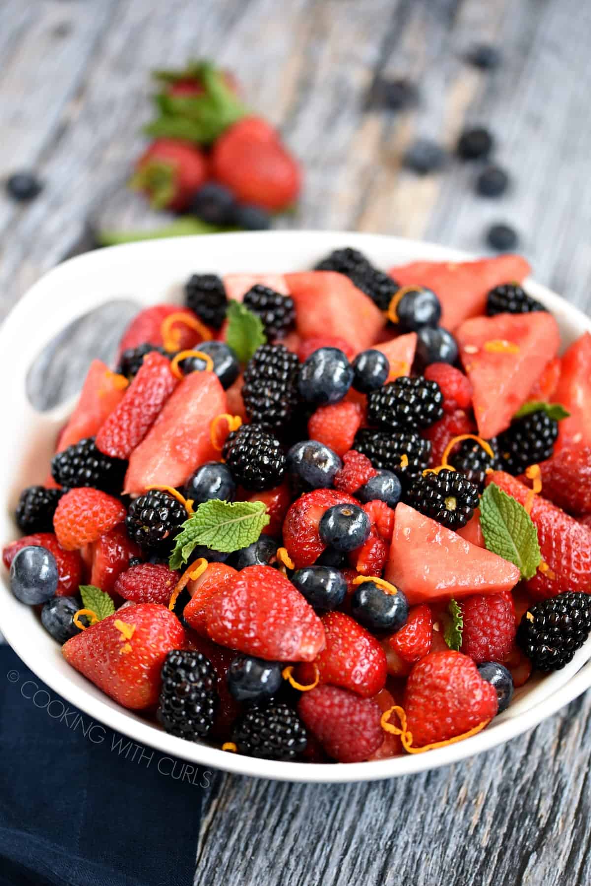 Watermelon wedges, blackberries, blueberries, raspberries and strawberry halves tossed together in a white serving bowl and garnished with mint leaves and orange zest.